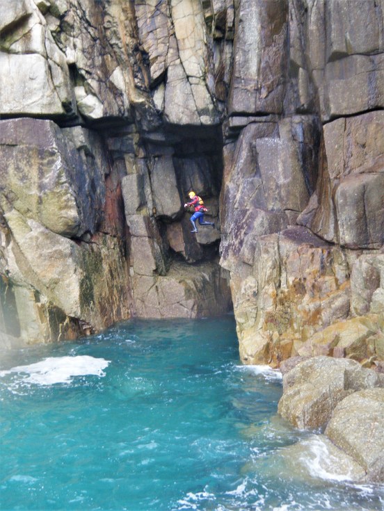 Coasteering guide doing a large cliff jump in front of a huge sea cave at Land's End