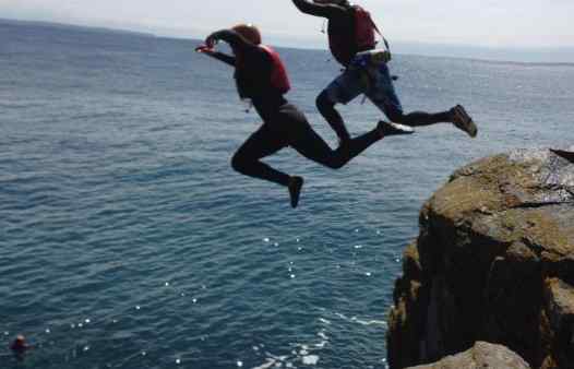 Two people coasteering, jumping off a large jump at Prussia Cove together.