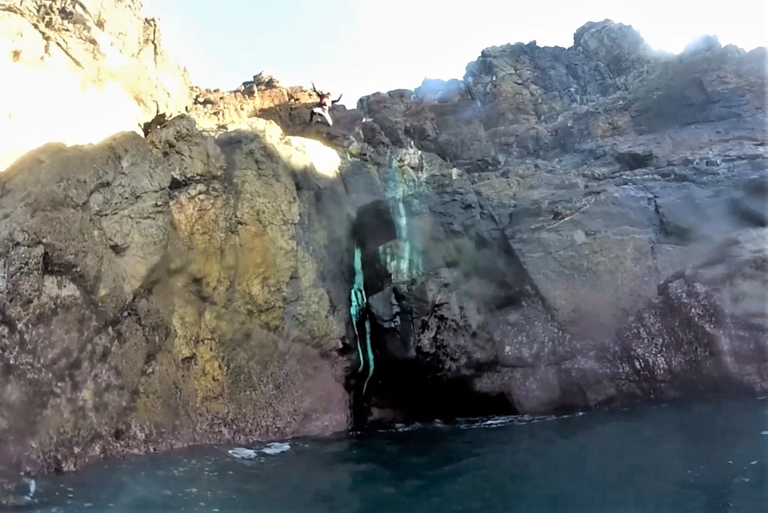 Coasteering guide jumping across the entrance of a small sea cave at Pendeen near Penzance, west Cornwall.