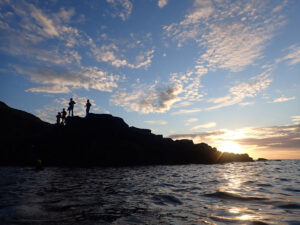 Group wearing coasteering equipment silhouetted at Land’s End at sunset