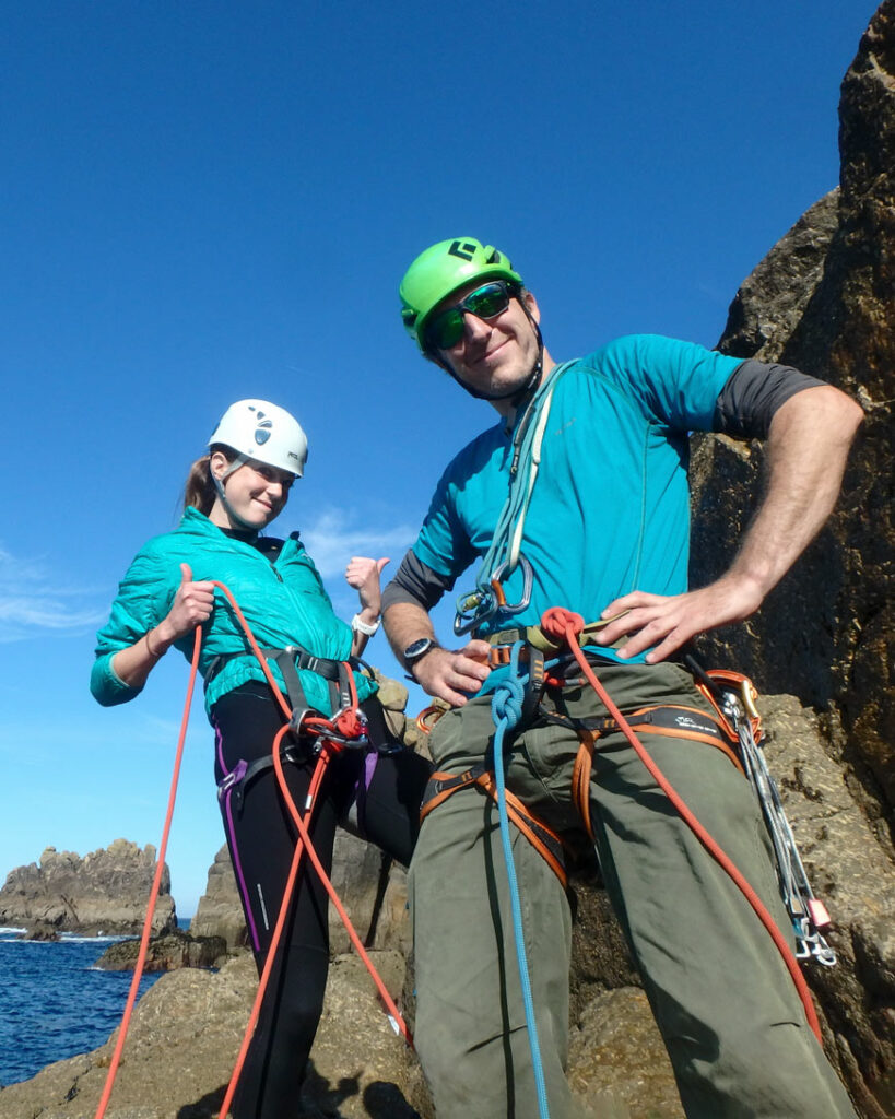 Two climbers in trad climbing harnesses at a sea cliff crag in Cornwall.