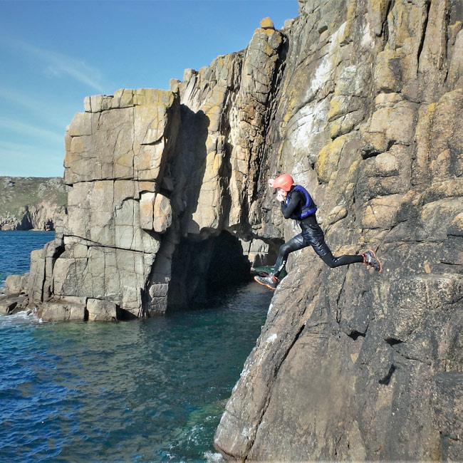 Adventurer leaps from exposed rock face beside a sea arch at Land’s End, Cornwall