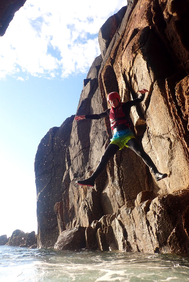 Coasteering participant edges along granite ledge above deep water before entering a dark Cornish sea cave
