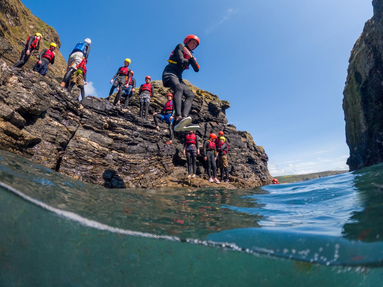 Young coasteer jumps into clear Cornish water as an experienced guide supports a family group nearby