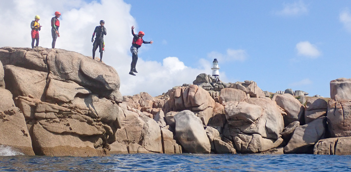 Man cliff jumping off a small granite island into the Atlantic with Peninnis Head Lighthouse in the background.