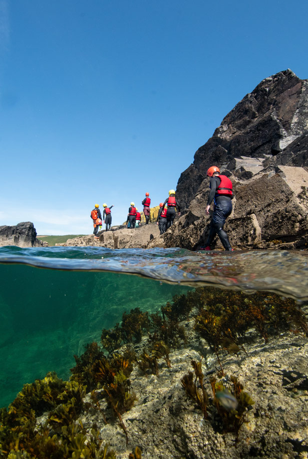 Coasteering group moving across intertidal rocks with seaweed visible underwater.