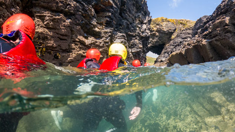 Group of coasteering participants swimming through a rock arch at Praa Sands, Cornwall