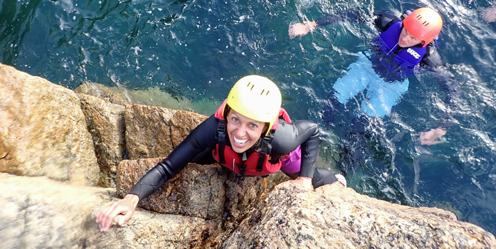 Smiling woman climbing out of the sea onto granite ledges during a Land’s End coasteering session