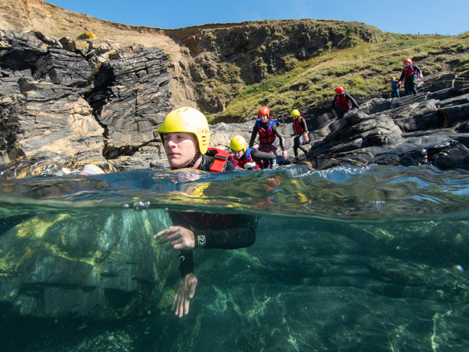 Coasteering participant in the water near rocky cliffs with a group preparing to jump.