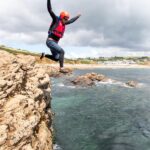 First-time coasteer leaps from a low cliff with Praa Sands beach in the distance