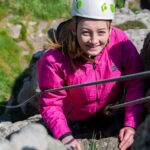 A young climber smiling confidently while scaling a rock face under supervision during a family-friendly climbing session