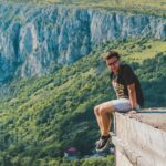 Man enjoys scenic mountain view sitting on cliff, capturing adventure and freedom.