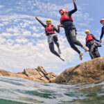 Group of smiling coasteering guides and adventurers jumping from granite rocks into the sea on the Cornish coast.