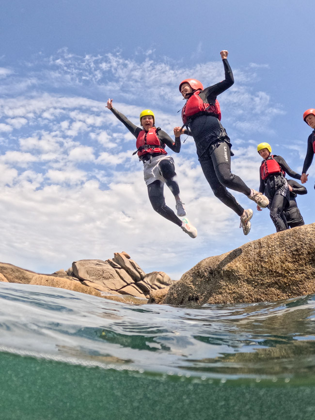 Group of smiling coasteering guides and adventurers jumping from granite rocks into the sea on the Cornish coast.